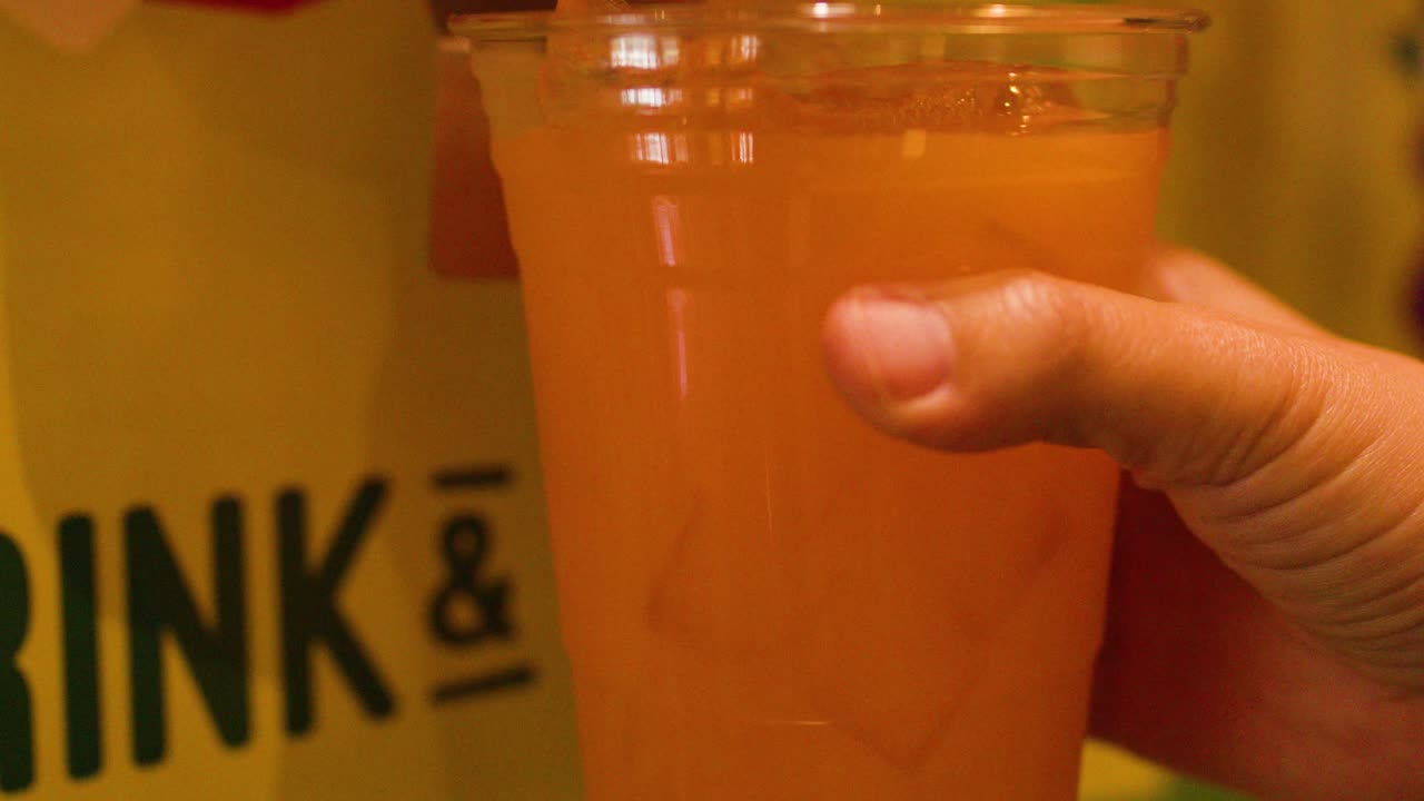 Hand fills plastic cup with iced orange beverage from push-button dispenser under warm indoor lighting