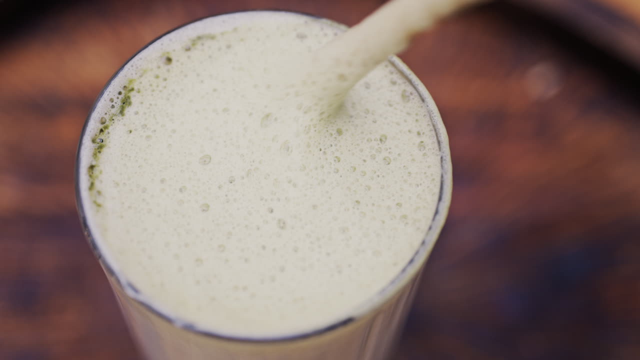 Close up of a woman mixing the foam on a matcha latte with a paper straw on a wooden tray