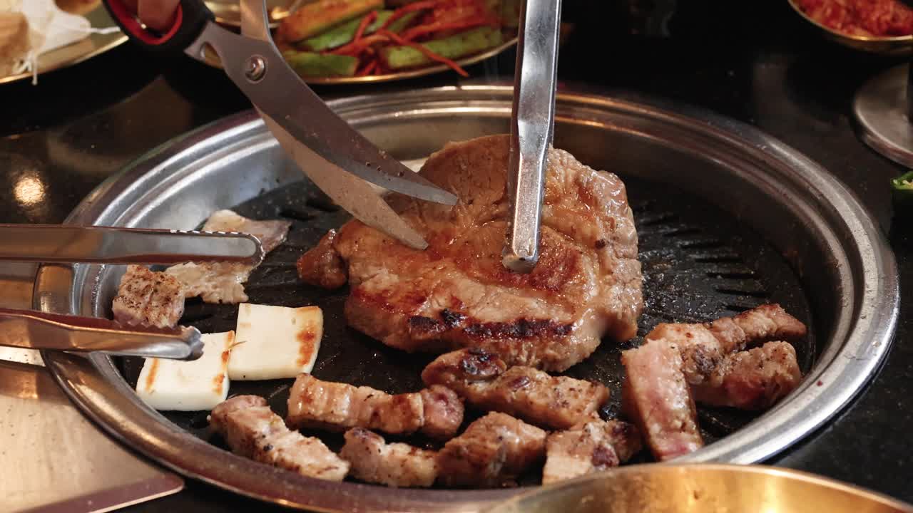Tongs and scissors cut pork belly on tabletop grill, bright lighting, close-up, indoor restaurant