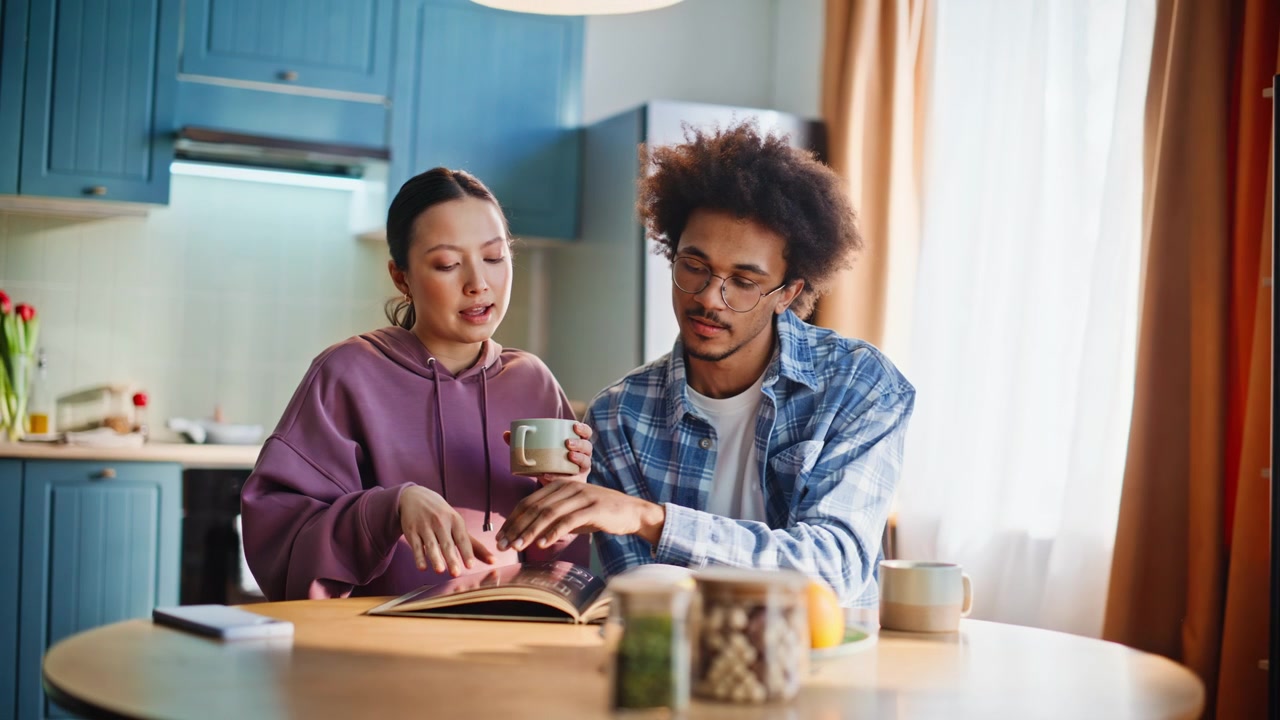 Multiethnic couple having breakfast at kitchen. Woman drinks coffee, man reads