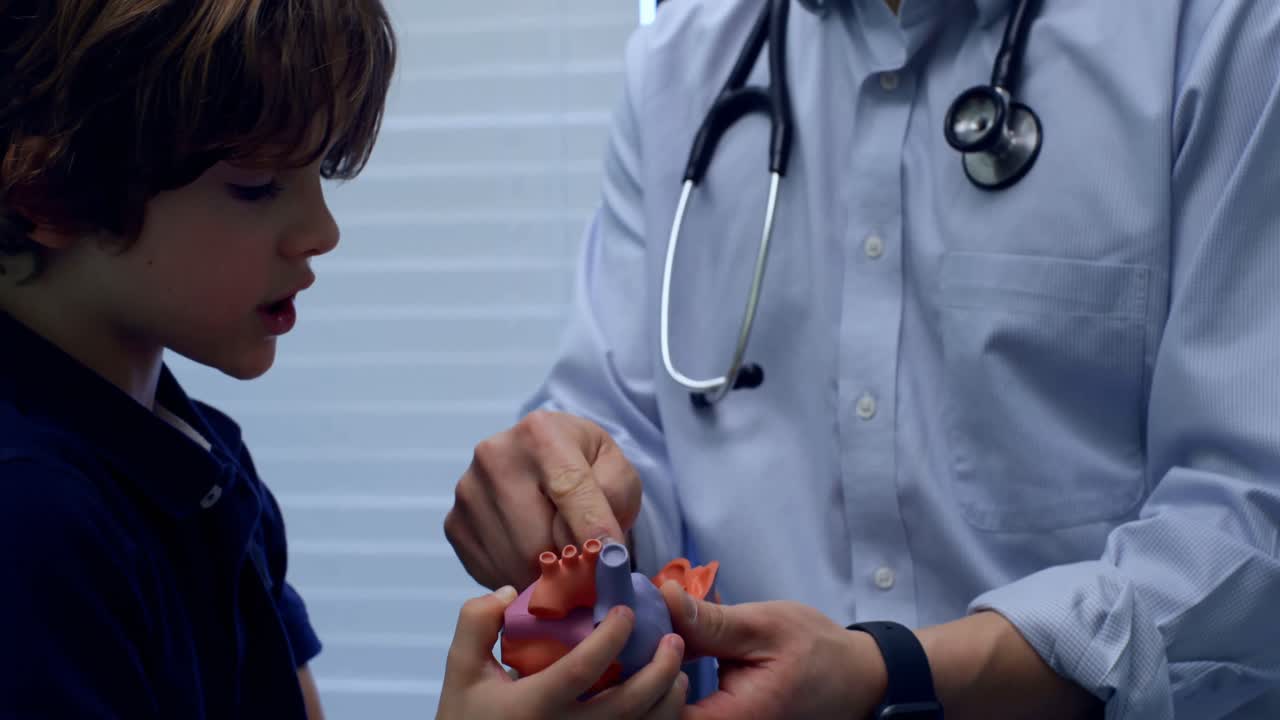 젊은 아시아 남성 의사가 클리닉 4k에서 백인 소년 환자에게 심장 모델을 보여주는 측면 뷰