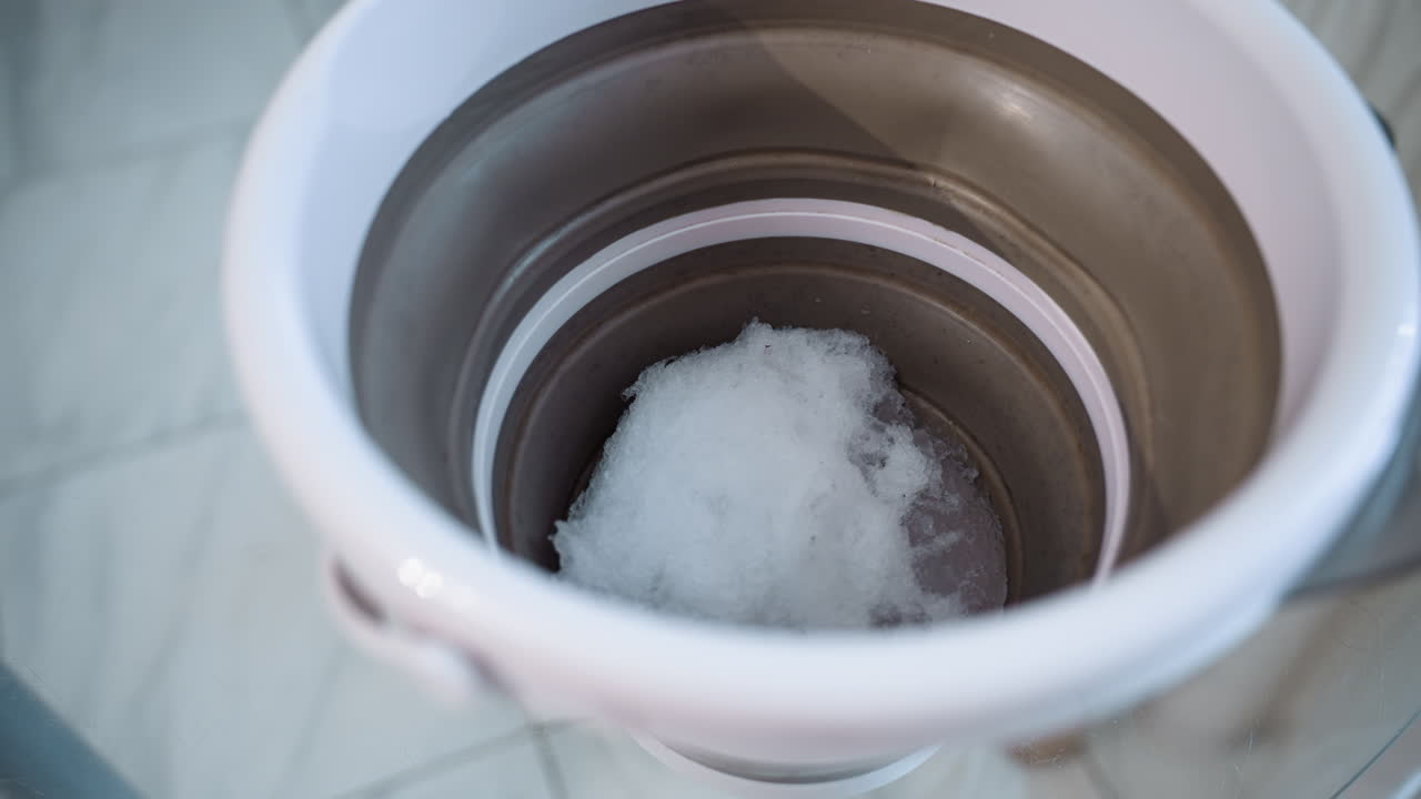 Bucket filled with fresh snow sits on plastic topped table under soft indoor light. Blurred background hints at home interior while subtle snow crystals and moisture droplets add delicate texture