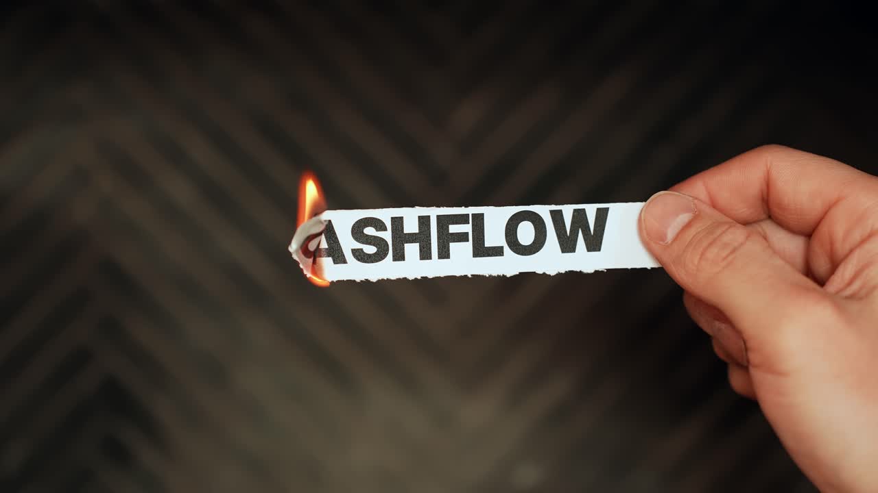 Close-up hand burns a paper label reading “CASHFLOW” with a lighter flame on dark background—finance stress, liquidity problems, budgeting crisis concept, copy space