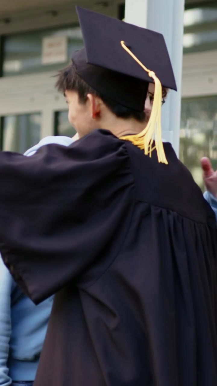Video vertikal: Pria berjalan mengenakan hoodie menggoyangkan, memeluk jumbai toga wisuda di pintu merayakan kelulusan