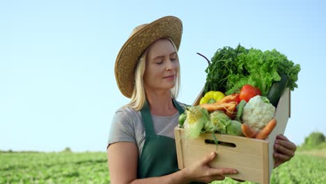 Portrait-Of-The-Blonde-Young-Pretty-Woman-Farmer-In-A-Hat-Demonstrating-A-Box-Full-Of-The-Mature-Vegetables-In-The-Green-Field