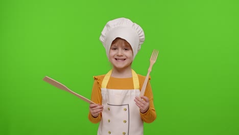 Child-girl-kid-dressed-as-cook-chef-showing-wooden-fork-and-spatula,-smiling,-nods-head-in-agreement