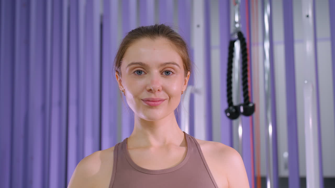 женщина с коричневыми волосами, привязанными назад, уверенно улыбается в камеру в тренажерном зале на фиолетовом фоне, энтузиаст фитнеса, излучающий позитивность и мотивацию после тренировки