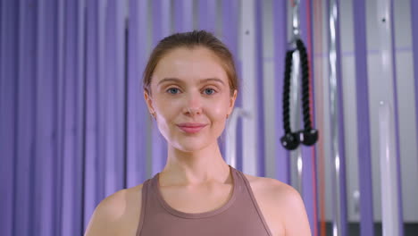 mujer con el cabello de sello marrón atado hacia atrás sonríe con confianza a la cámara en un entorno de gimnasio con un fondo púrpura, entusiasta del fitness que irradia positividad y motivación después de una sesión de entrenamiento