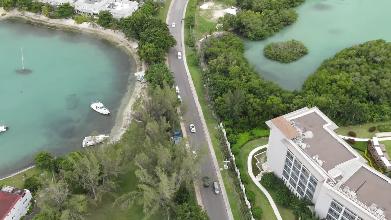 Drone footage capturing a sleek car cruising through the upscale community of Freeport, Montego Bay, Jamaica. Lush greenery, ocean views, and luxury villas line the scenic, well-paved road