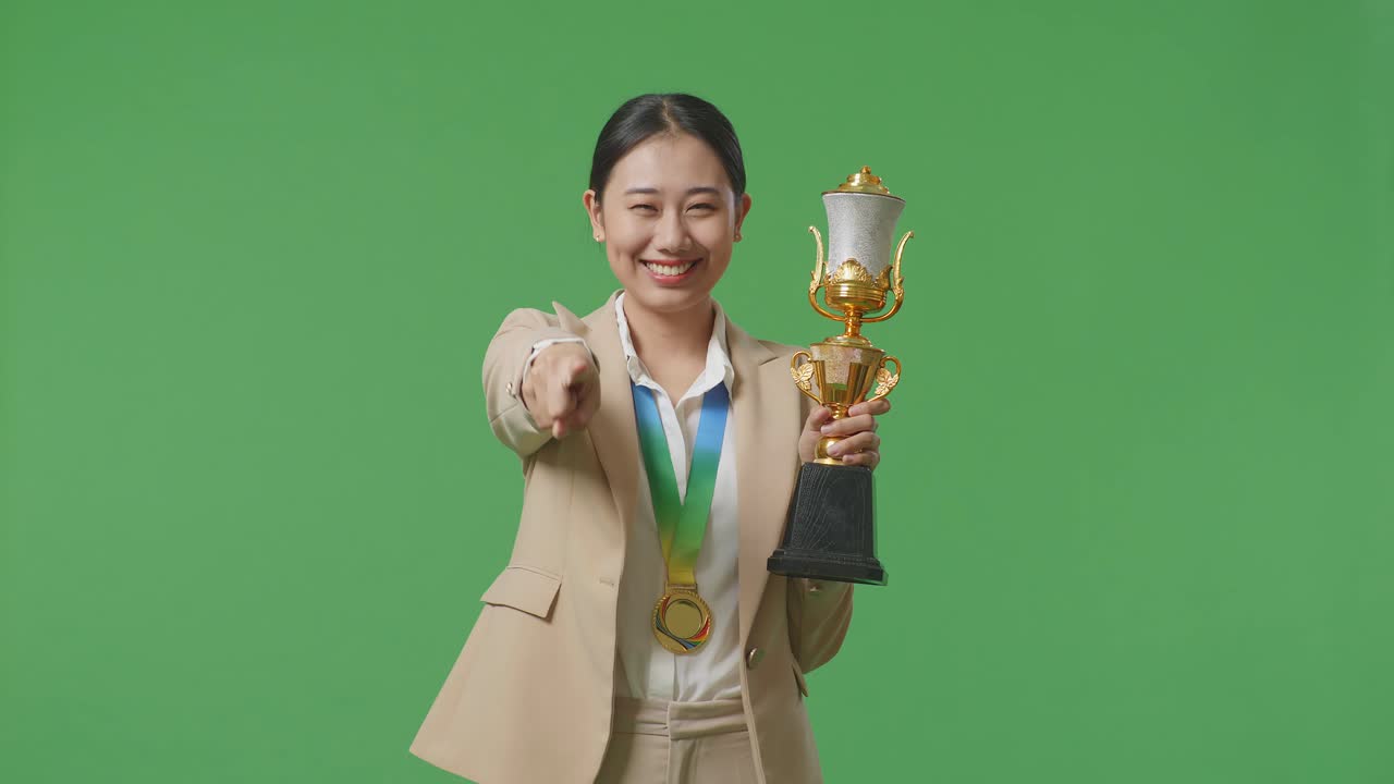 금메달과 트로피를 입은 슈트를 입은 아시아 사업가 여성이 가을 만지고 미소 짓고 카메라를 가리키며 스튜디오의 녹색 화면 배경에서 자랑스러워합니다.