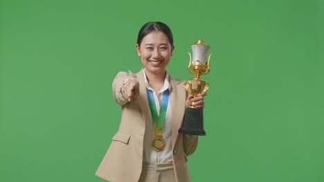mujer de negocios asiática en traje con una medalla de oro y un trofeo tocando su pecho luego sonriendo y señalando a la cámara estando orgullosa de sí misma en el fondo de la pantalla verde en el estudio