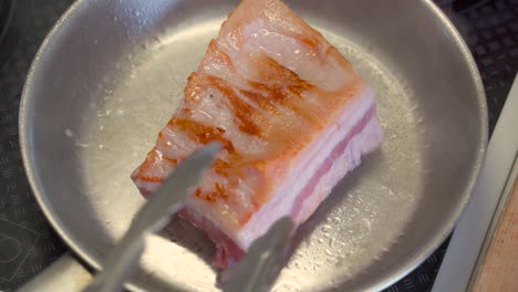 top down view of pork belly being cooked inside stainless steel pan, moved around with tongs