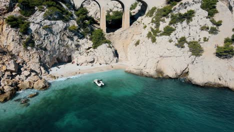 Aerial-pullback-from-boat-reveals-picturesque-bridge-arch-in-coastal-region-of-France,-green-shrubs-dot-landscape