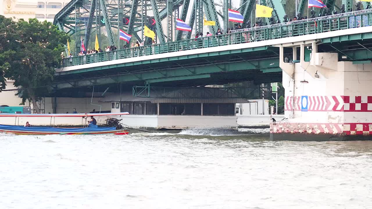 A boat travels under a bridge in Bangkok, Thailand. Bright daylight, steady camera, and urban river setting
