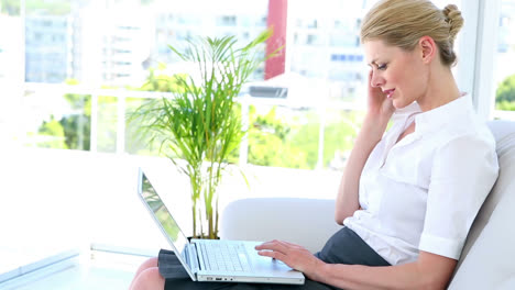 Businesswoman-sitting-on-couch-using-laptop-talking-on-phone