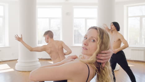 Portrait-Of-A-Blonde-Female-Contemporary-Dancer-Looking-At-Camera-And-Training-Dance-Moves-With-Two-Men-In-The-Studio