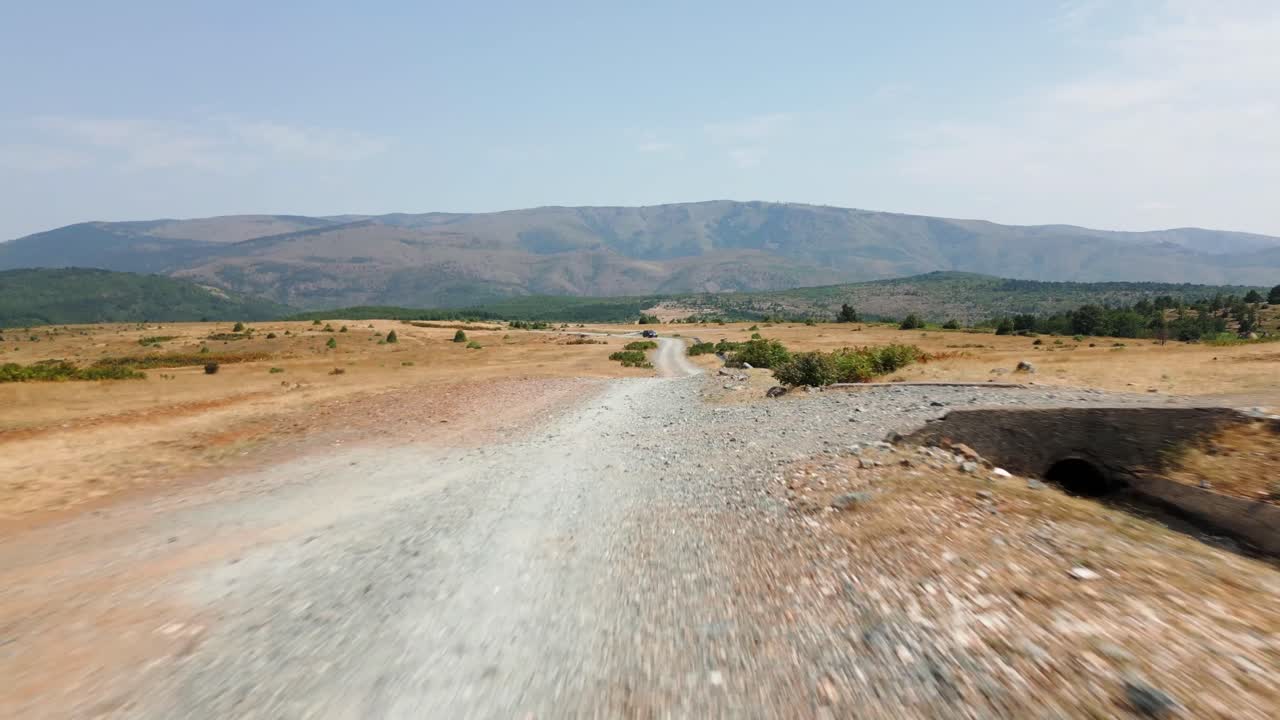 Car driving through dry countryside with mountains in the background on a sunny summer day in Albania. Scenic road trip and travel adventure footage
