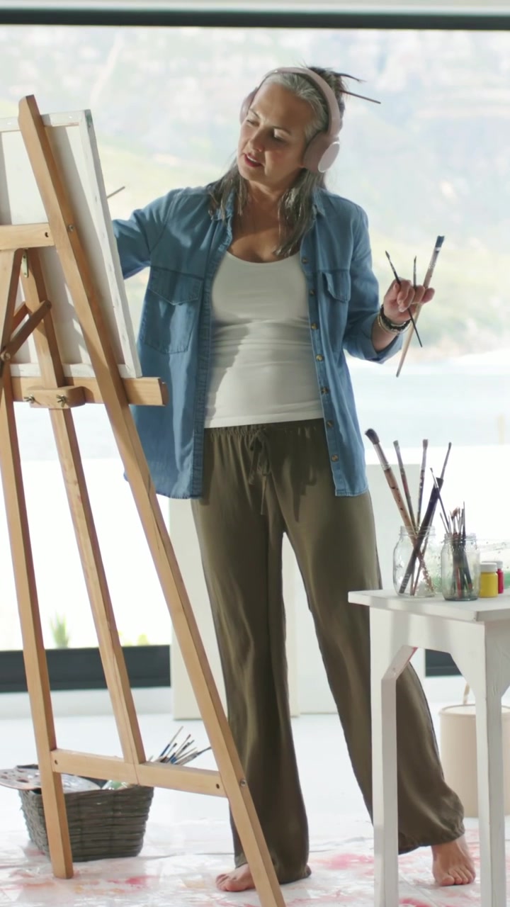 Video verticale: Barattoli in primo piano, donna in camicia che prende un pennello e dipinge su un cavalletto in studio.