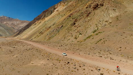 cyclist and suv on dirt road in arid cajón del maipo canyon, adventure travel and mountain biking in andes mountains, chile. aerial backward