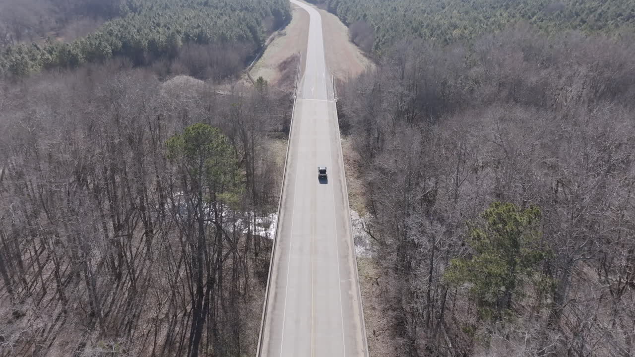 Aerial footage of a lone vehicle drives down a straight, elevated road cutting through a dense forest of leafless trees, with a small creek visible below.