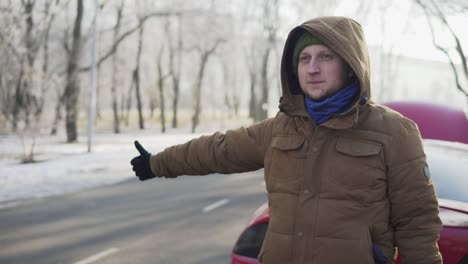 Attractive-young-man-hitchhiking-on-a-road-with-his-broken-down-car-behind-him