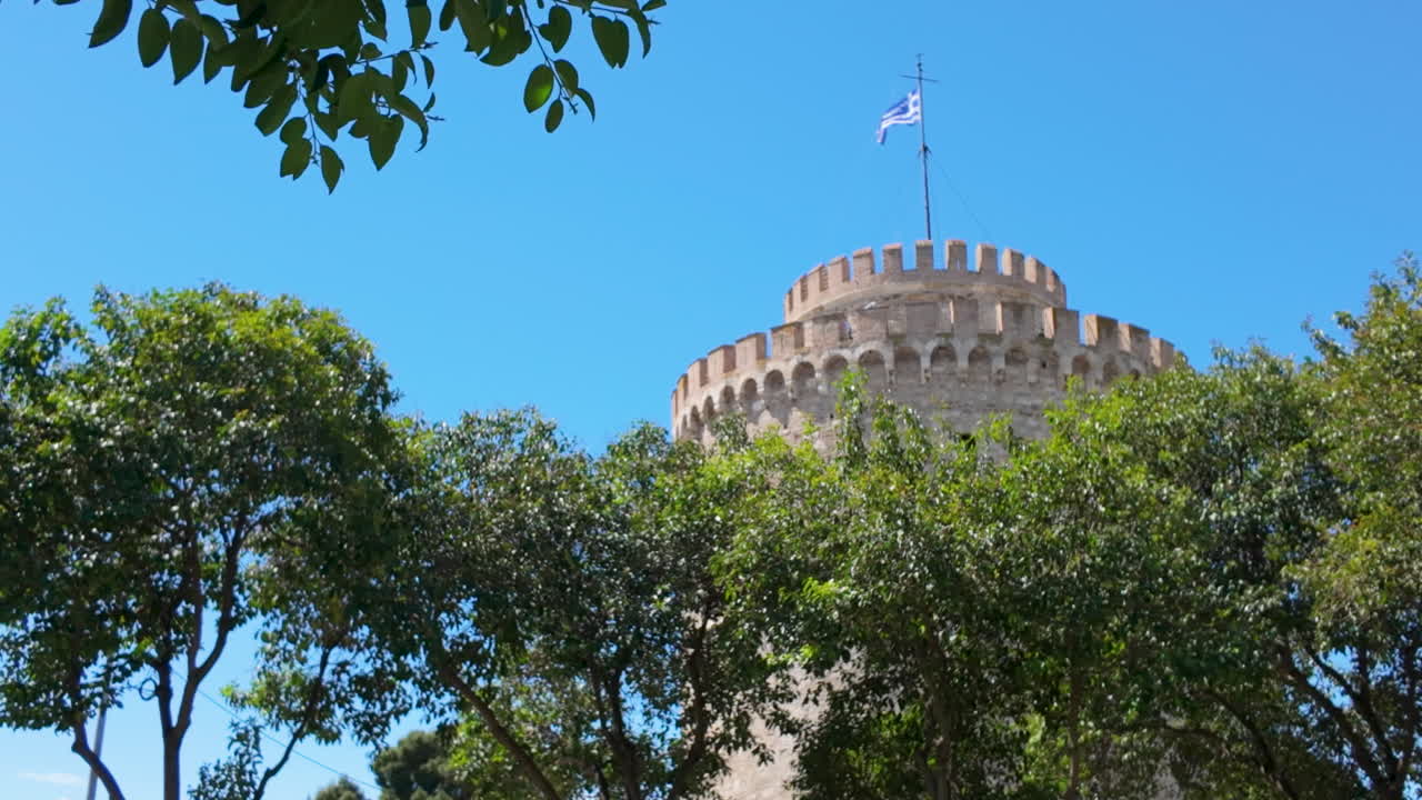 Una famosa torre rotonda in pietra si erge sopra lussureggianti alberi verdi sotto un cielo azzurro in Grecia, con una bandiera che sventola sulla cima