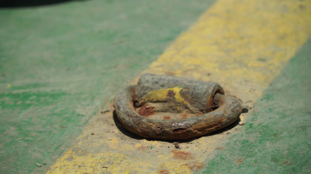 Anello di ancoraggio per parcheggio vicino a striscia gialla, metallo incassato usurato che mostra corrosione e vernice scheggiata su corsia di cemento in un angolo di garage illuminato dal sole con superficie granulosa, utile per la sicurezza, infrastruttura.