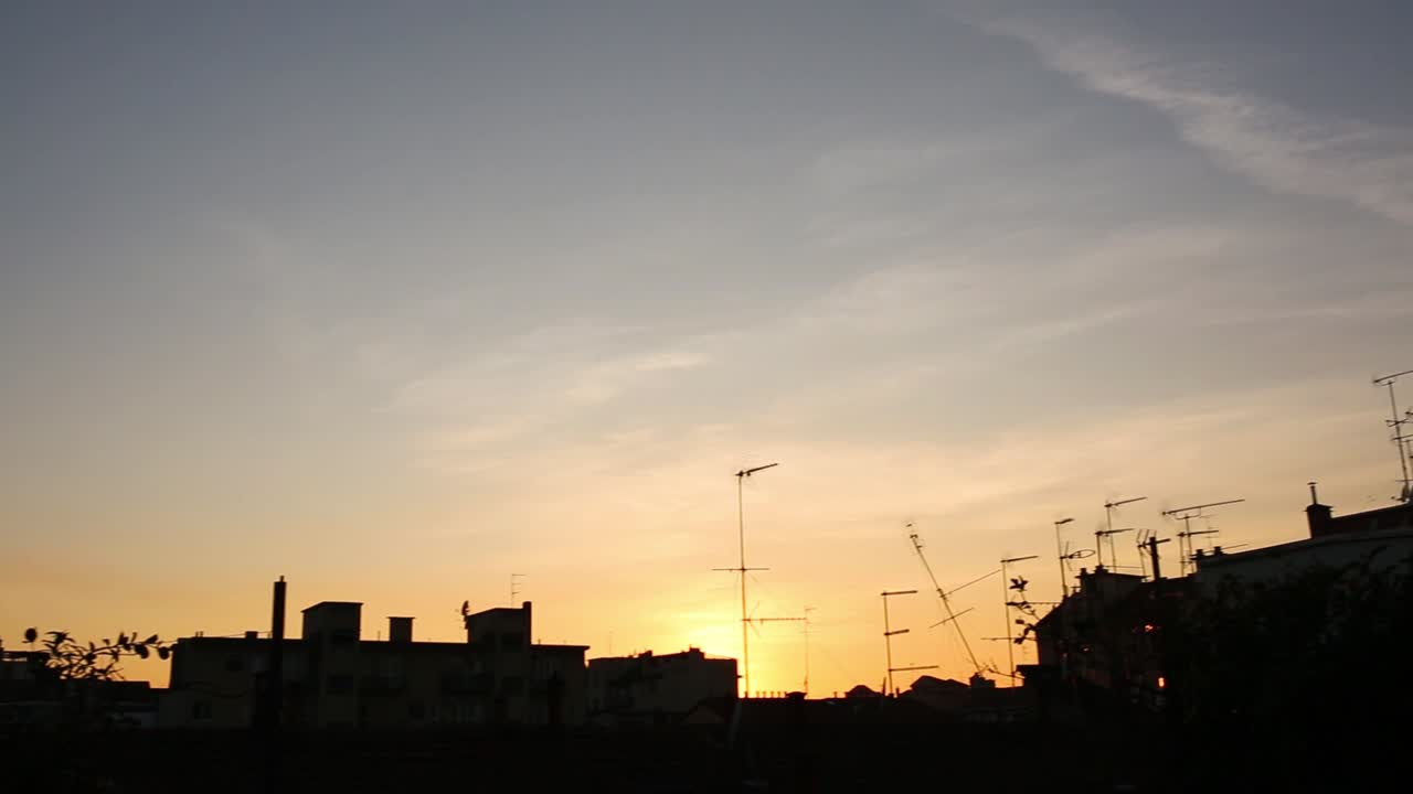 movimiento panorámico de la cámara en el balcón, vista de los edificios de los tejados en silueta y puesta de sol naranja y cielo azul nublado en el fondo