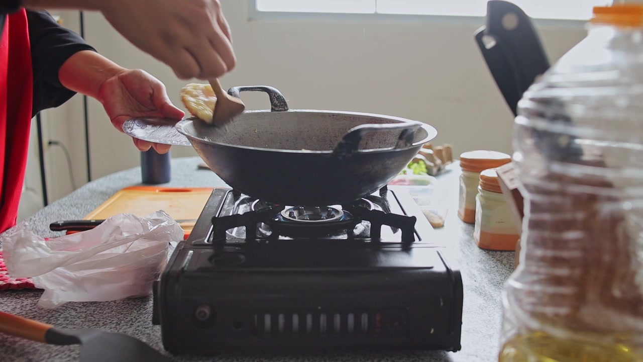 Koki menggunakan spatula kayu untuk mengaduk bahan-bahan di wajan panas di atas kompor gas portabel di dapur rumah