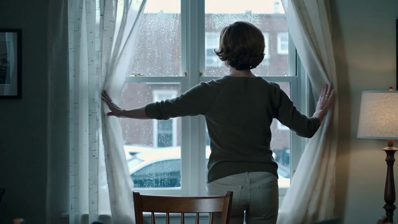 Flipping notebook, woman in olive sweater at home table parting curtains to view rainy street