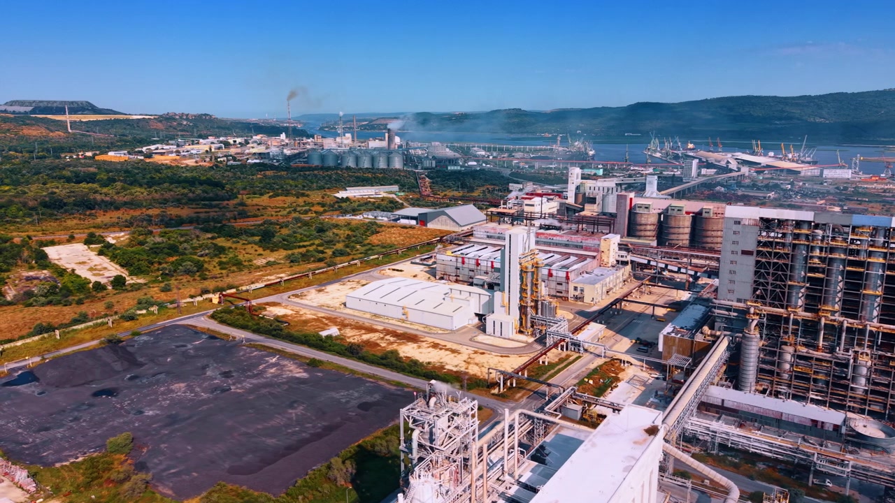 Edificios de fábrica y sistemas de tuberías cerca de un puerto de carga. Una vista de dron captura estructuras de producción, carreteras y vegetación con silos de almacenamiento y grúas portuarias a lo lejos.