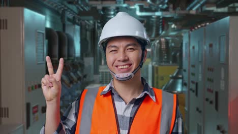 close up of asian male engineer with safety helmet smiling and showing peace gesture while standing in engine control room, work of electrical generators