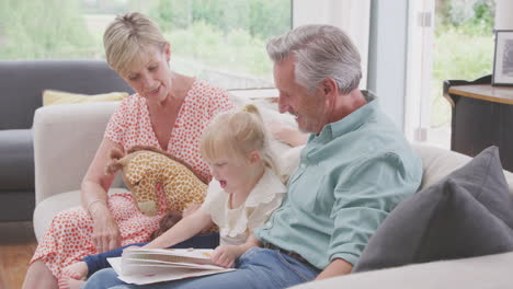 Abuelos-Sentados-En-El-Sofá-Con-Su-Nieta-En-Casa-Leyendo-Un-Libro-Juntos