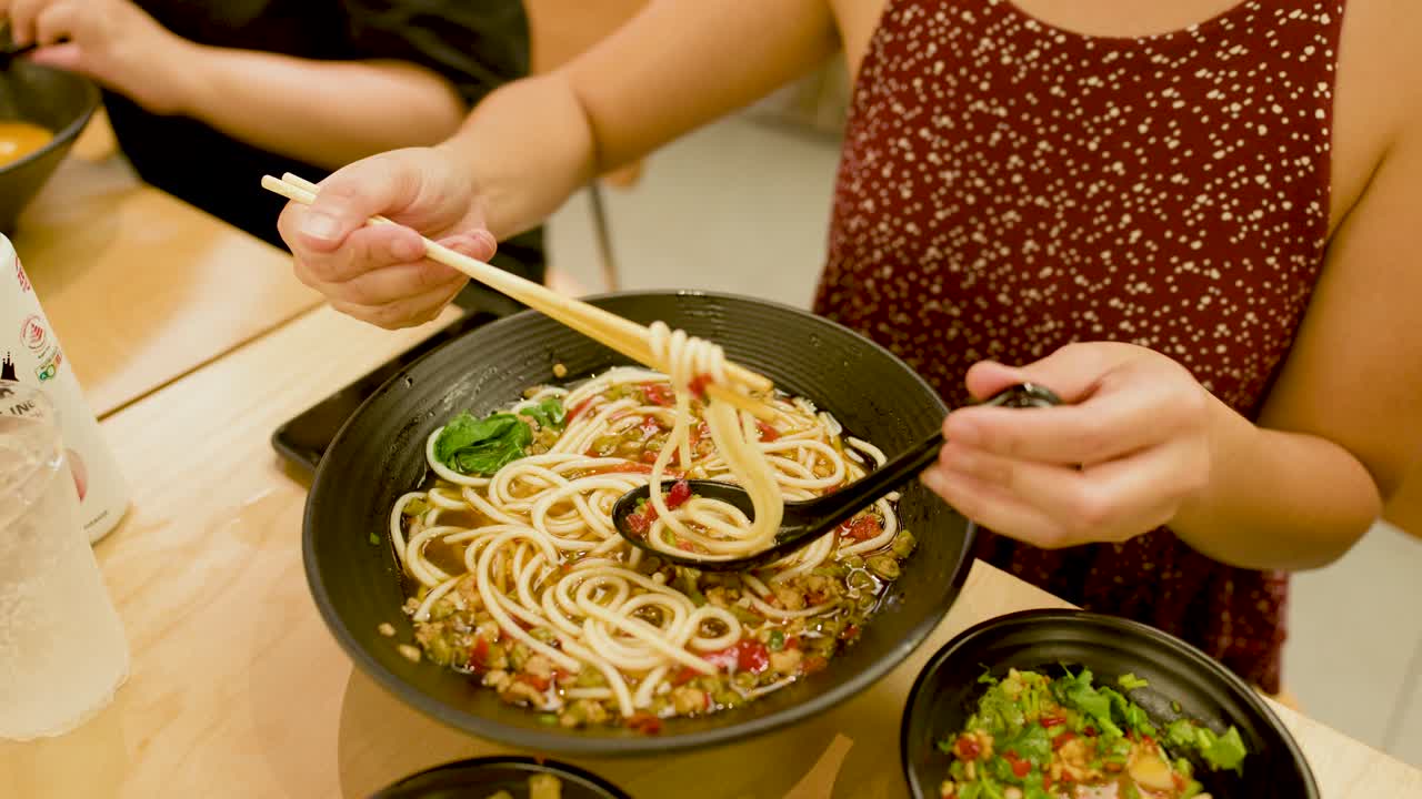 Woman uses chopsticks and spoon to lift spicy noodle soup in bright, casual dining setting