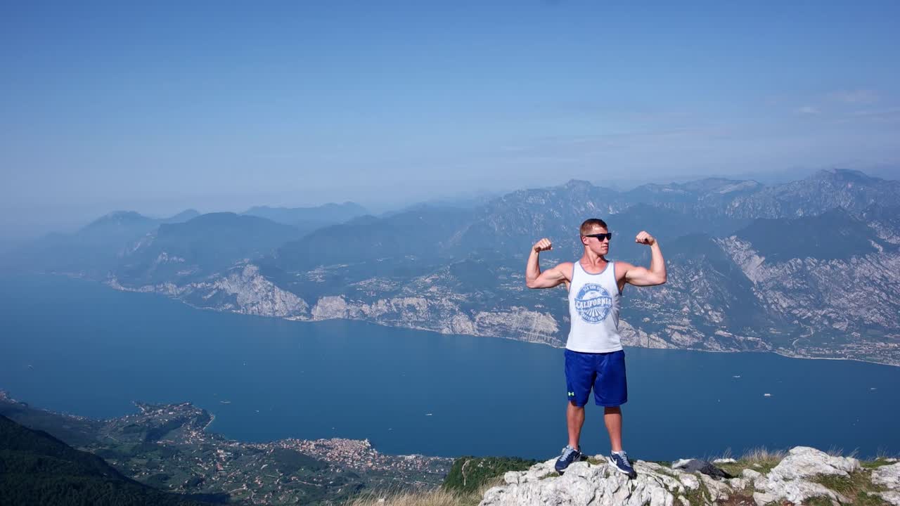 A man posing at the top of the hill on Lake Garda, Italy. The male is flexing his muscles as the camera pans from left to right with part of the lake and surrounding mountains and hills on display.