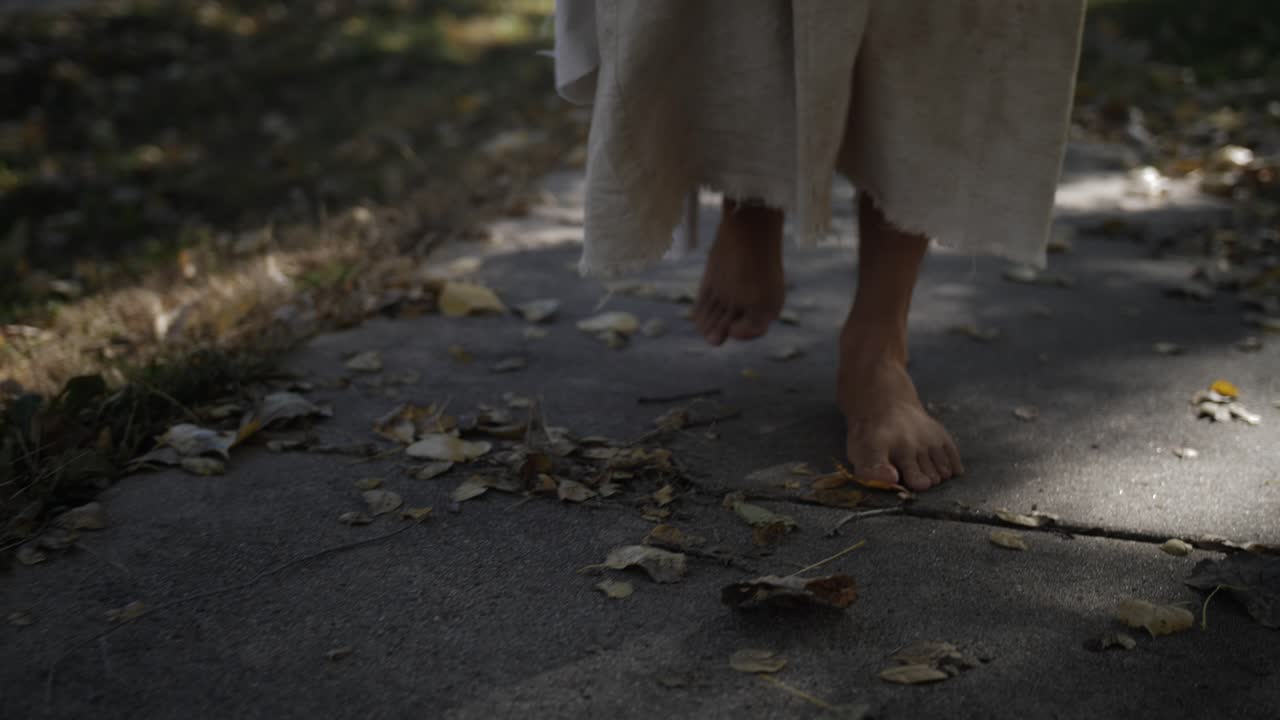 A closeup shot of a man dressed as Jesus Christ wearing a white, tattered robe walks in dramatic slow motion on a sidewalk