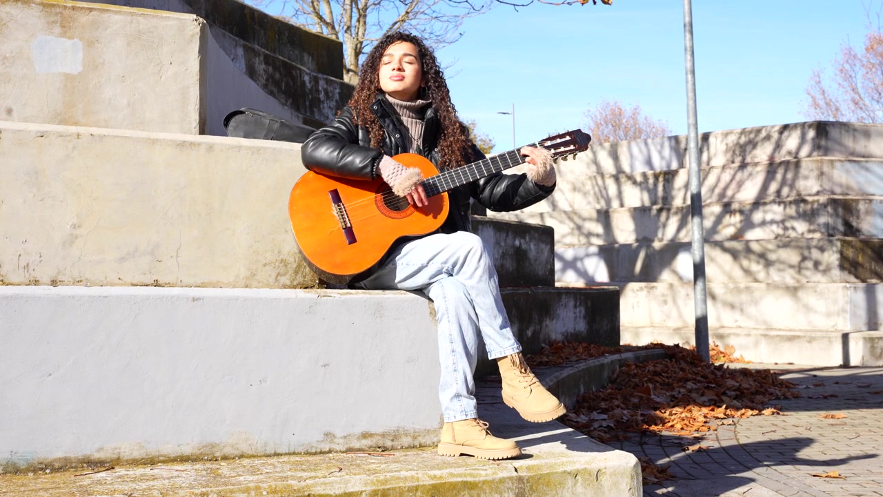 En kvinde med krøllet hår spiller akustisk guitar, siddende på betontrapper i en park på en solrig efterårsdag.