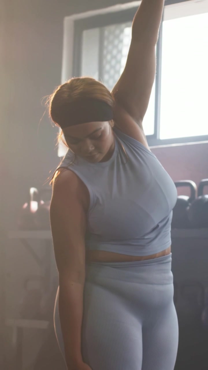 Video verticale: Donna in completo blu posiziona i piedi vicino ai manubri, si riscalda con uno stretching laterale in palestra.