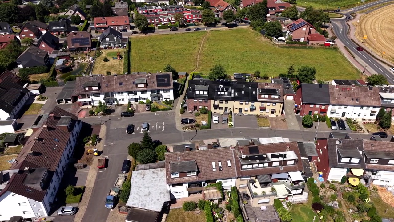 Reihen von Reihenhäusern mit Solardächern säumen ruhige Wohnstraßen in einer kleinen deutschen Stadt, grüne Felder und goldene Weizenfelder umgeben das Viertel darunter. Luftaufnahme mit Drohne, seitliche Weitwinkelaufnahme
