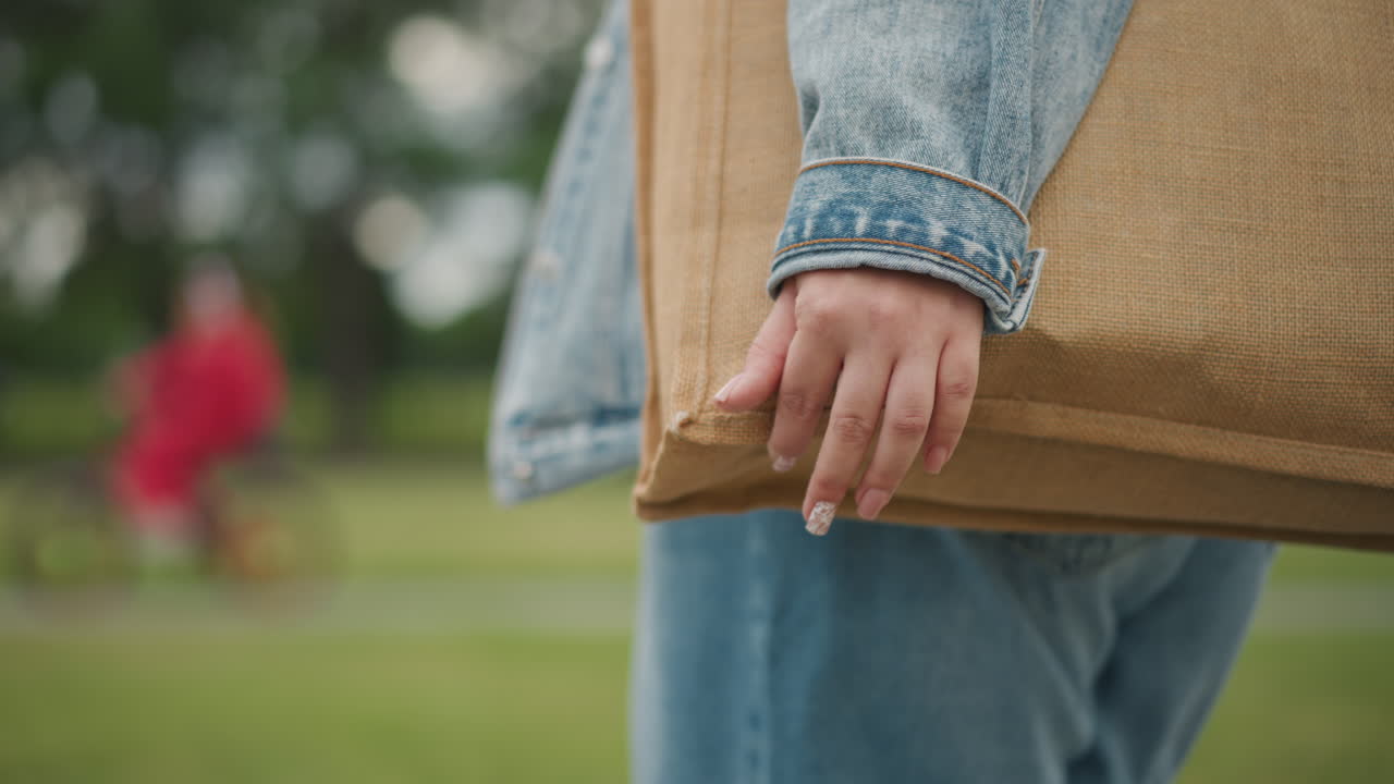 Nærbilde av hånd som holder en vevd veske, dongerijakke, manikyrerte negler, uskarp syklist i grønn park, avslappet sommerpromenade, teksturert veskedetalj, uformell helgemarkedsstemning, miljøvennlig shopping i naturlig dagslys