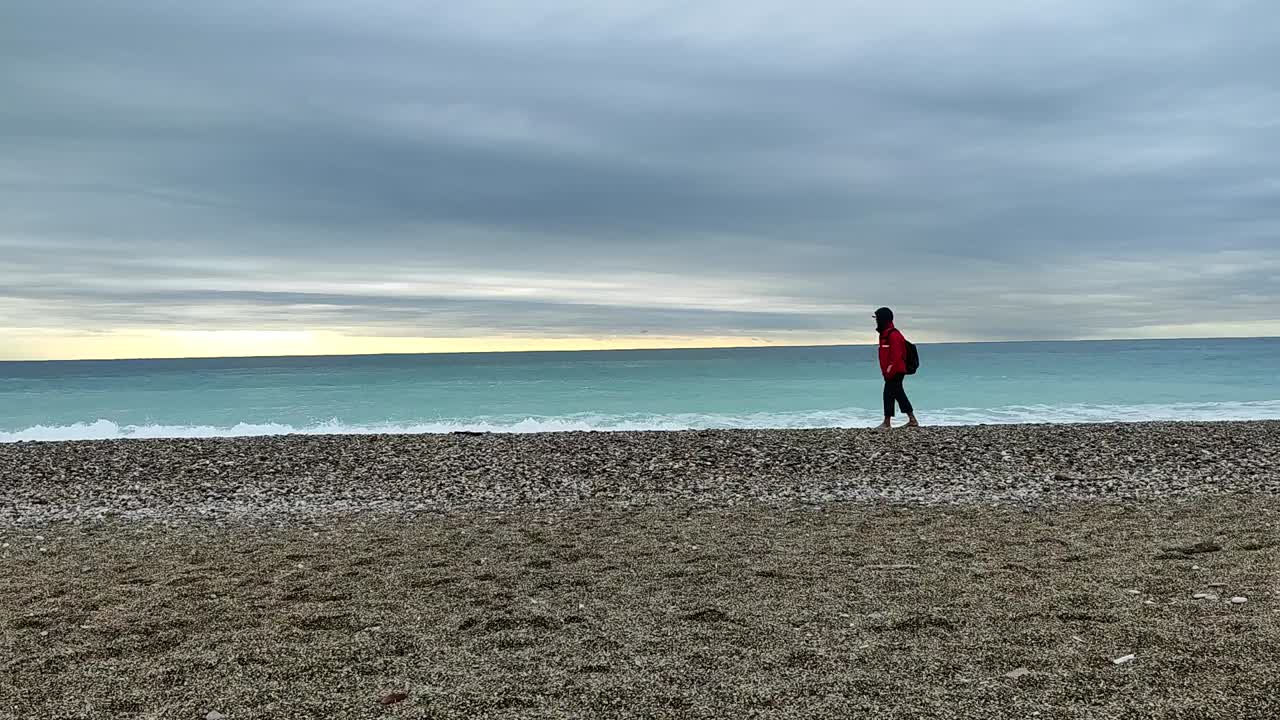Person in red jacket stands alone on pebbled Konyaaltı Beach, gazing at turquoise waves under an expansive overcast sky. Captured in Antalya, Turkey, the serene setting evokes tranquility