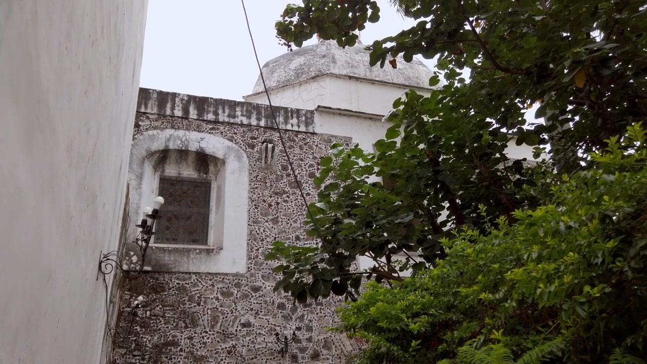 Exterior de iglesia colonial con muros de piedra desgastados, techo abovedado, ventana arqueada, adorno ornamentado y exuberante follaje rodeando el edificio histórico en México