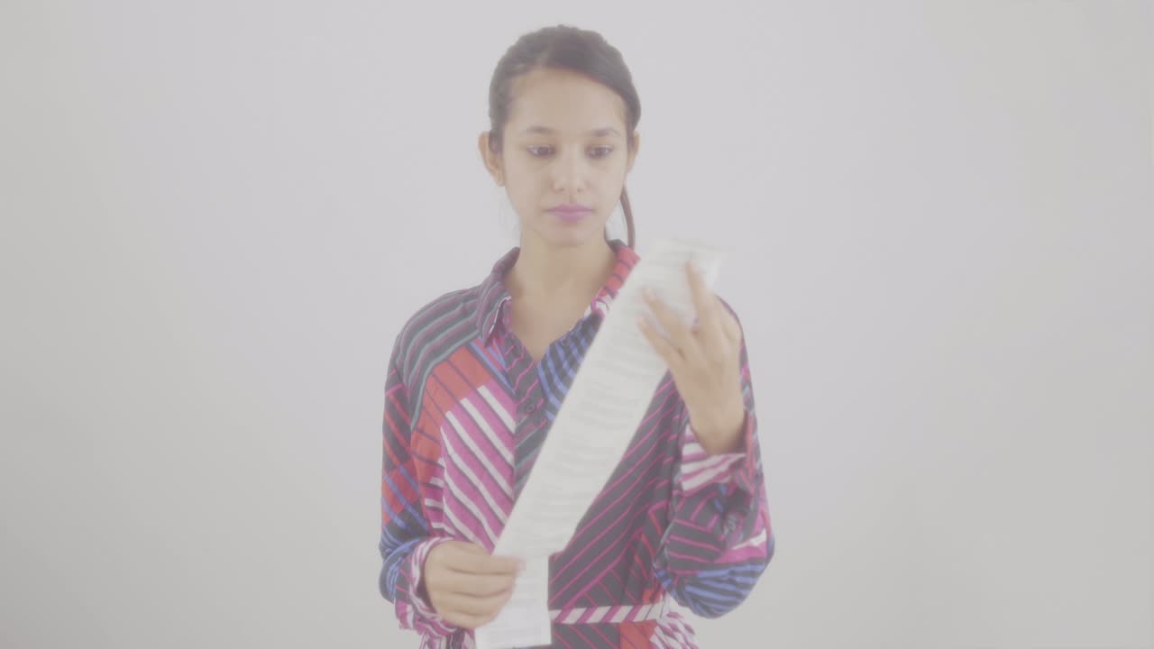 긴 종이 청구서 영수증을 보는 소녀는 비용을 계산하고 가격을 확인하고 카메라를 보고 충격을 받았습니다.