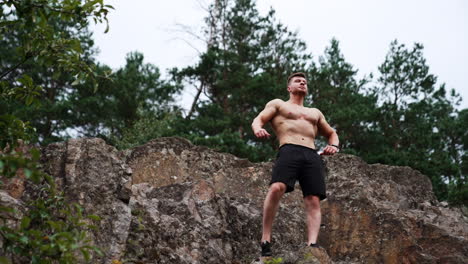 athletic man with naked torso wearing black shorts stands on the rock. sportsman rises hands showing muscles. low angle view.