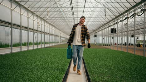 portrait of a confident guy in a farmer cap walking along plant sprouts and carrying a blue watering can in his hands while working in a greenhouse on a farm