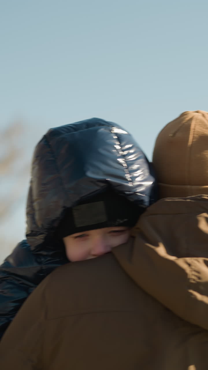 un primer plano de un padre llevando a su hijo y caminando al aire libre, el padre está vestido con una chaqueta marrón y gorra, y el niño en una chaqueta negra hinchada, con una vista borrosa de un edificio