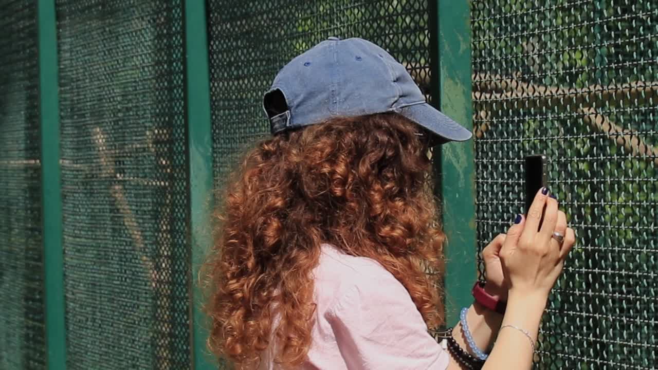 Person with curly hair and cap taking a photo through a fence