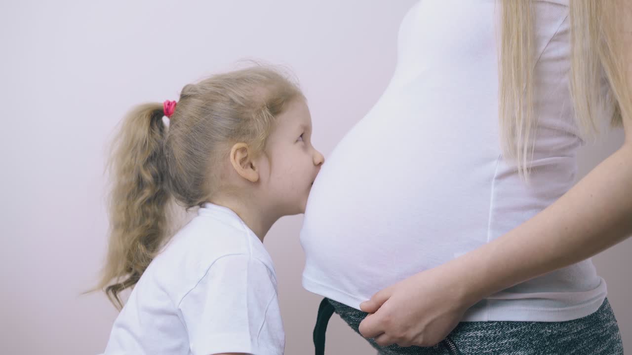 재미있는 소녀가 벽에 임신 한 엄마의 큰 배를 키스합니다.