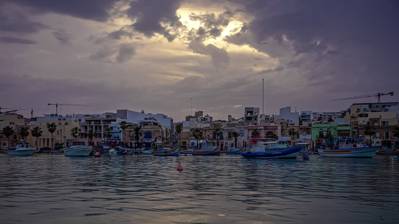 barcos blancos azules anclados en la ciudad portuaria mientras el sol se pone sobre marsaxlokk, malta, timelapse