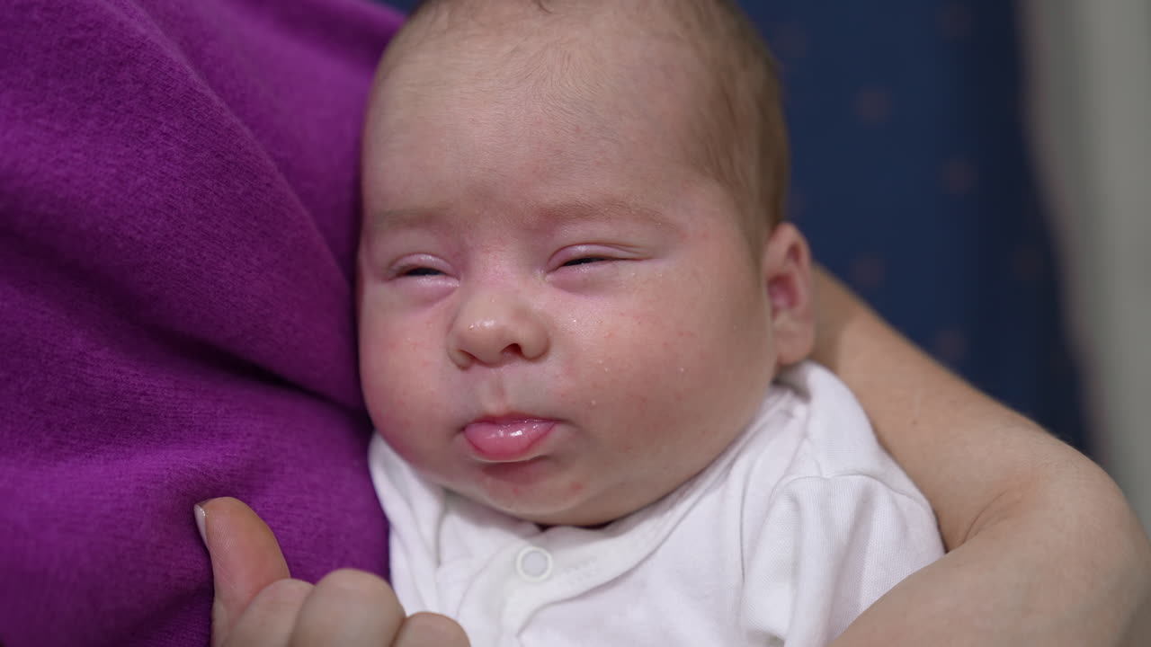 Baby doesn't want to wake up and barely opens his eyes. Mother is touching baby's nose gently to wake him up. Close up.